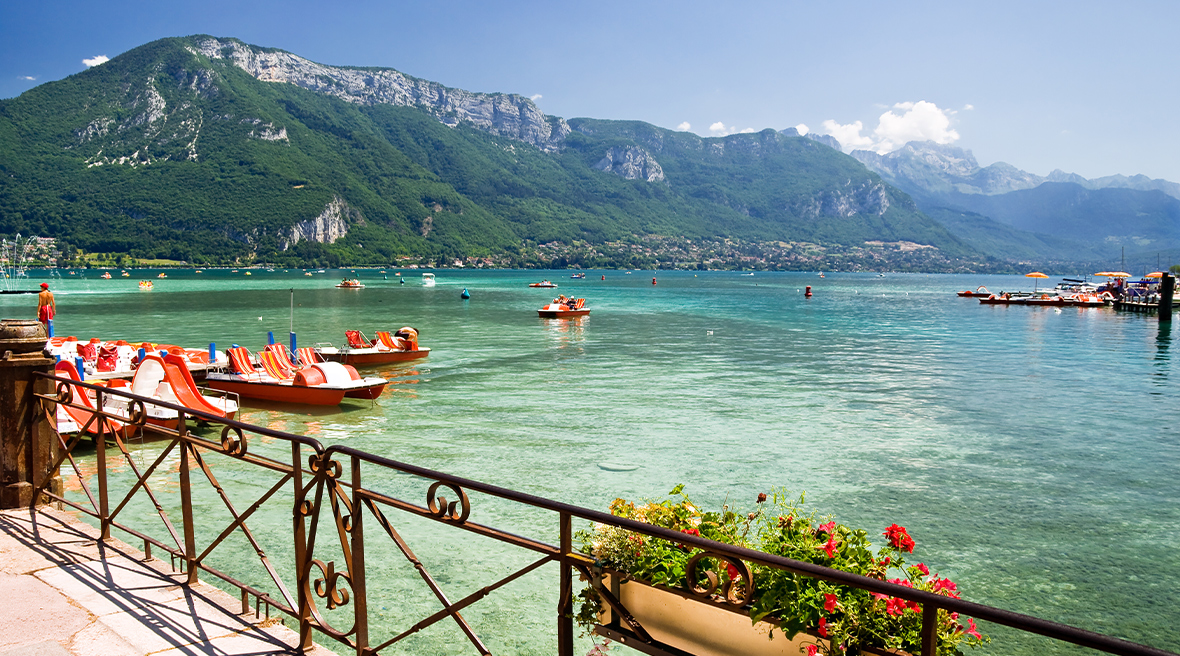 View of a lake with beautiful clear waters with a mountain range beyond. Pedalo boats on the lake and red flowers in a tub on the railings